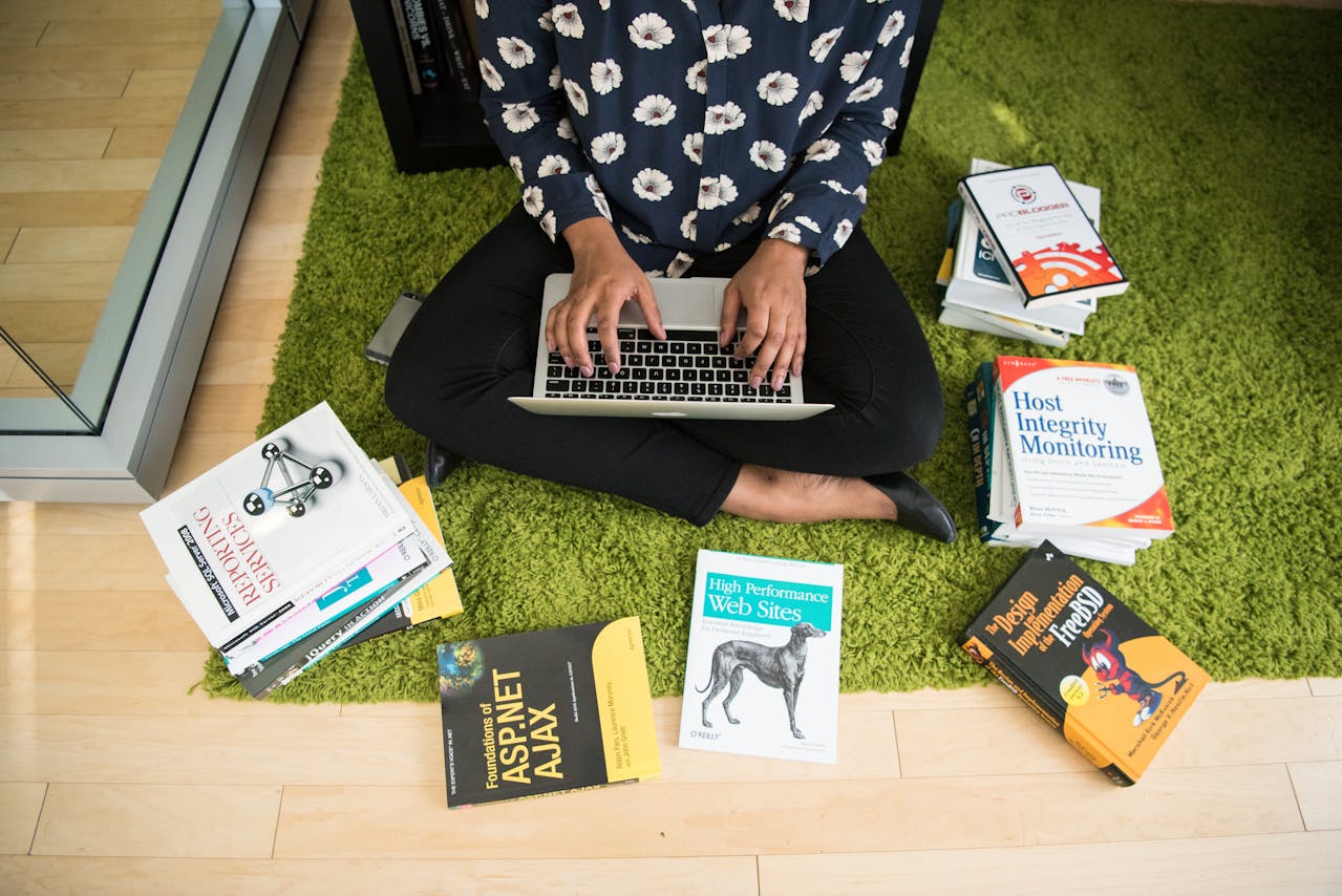 woman sits on floor with laptop, surrounded by books