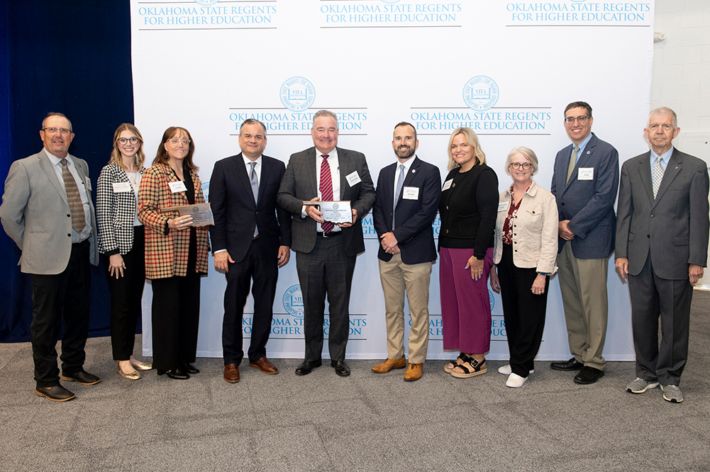 Ten people in business attire stand in front of a blue and white printed backdrop, holding two plaques in the shape of Oklahoma