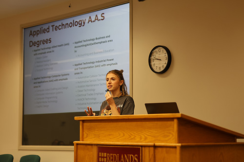 Woman standing behind a podium speaking into a microphone with a list of applied technology degree options on the screen behind her