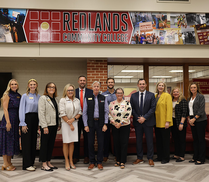 a group of men and woman standing in front of a photo collage of students and the logo for Redlands Community College