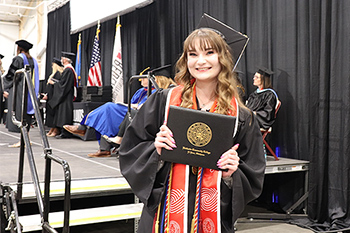 Smiling graduate with a red stohl and multiple colored cords holds her diploma case