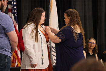 Woman in dark blue shirt applies a nursing pin to the white coat of a second woman