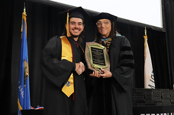 Two peope in graduation robes shake hands while holding a plaque