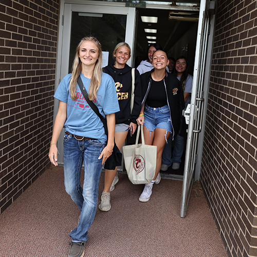 Female students wearing Redlands shirts are walking out of the door of a campus building during a tour.