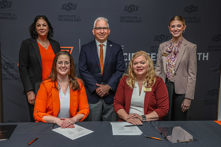 Representatives from both institutions are standing and sitting front of a dark wall. Participating in the signing are front row left to right Heather Ortiz, interim dean for the School of Arts, Sciences & Health, the School of Creative & Information Technologies, and the School of Engineering & Construction Technologies at OSUIT, and Annie Pearson, vice president of Academic Affairs at Redlands, and back row left to right Melissa Dreyer, assistant dean in OSUIT’s School of Arts, Sciences & Health; Chris Benge, OSUIT vice president of Operations; and Macey Scotten, coordinator of Strategic Partnerships at Redlands.