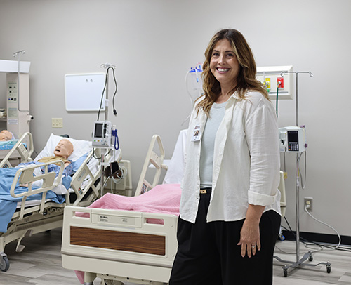 Tara Thomas is standing in front of hospital beds and simulation dummies in the nursing lab.