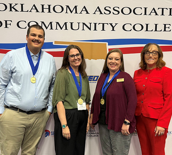 One man and three women wearing medallion awards stand in front of the Oklahoma Association of Community Colleges backdrop