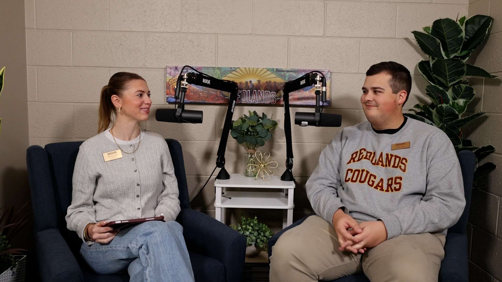Woman in gray sweater smiles at man in Redlands Cougars sweatshirt while sitting at podcast microphones