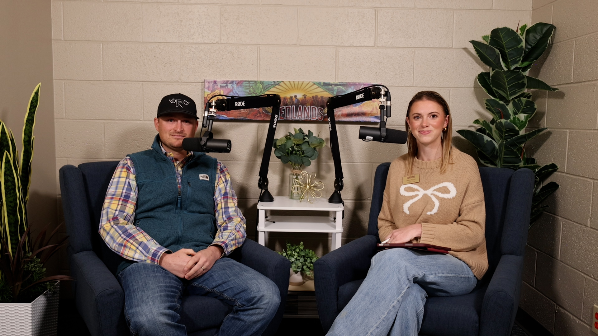 Man in multicolored plaid shirt and navy vest sits next to woman in tan sweater with cream bow stitched in