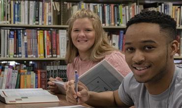 Two students smile at the camera while surrounded by books, and writing on paper
